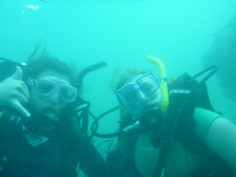 Two scuba divers are underwater, wearing masks and scuba gear. The diver on the left is giving a thumbs-up. The water is clear but slightly murky, with a greenish-blue tint. The divers appear to be exploring the underwater environment, possibly near a reef or other interesting underwater feature. The image captures the adventure and excitement of scuba diving.